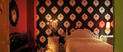 A bedroom with a patterned wallpaper, a bed, a nightstand with a lamp, and a chair.
