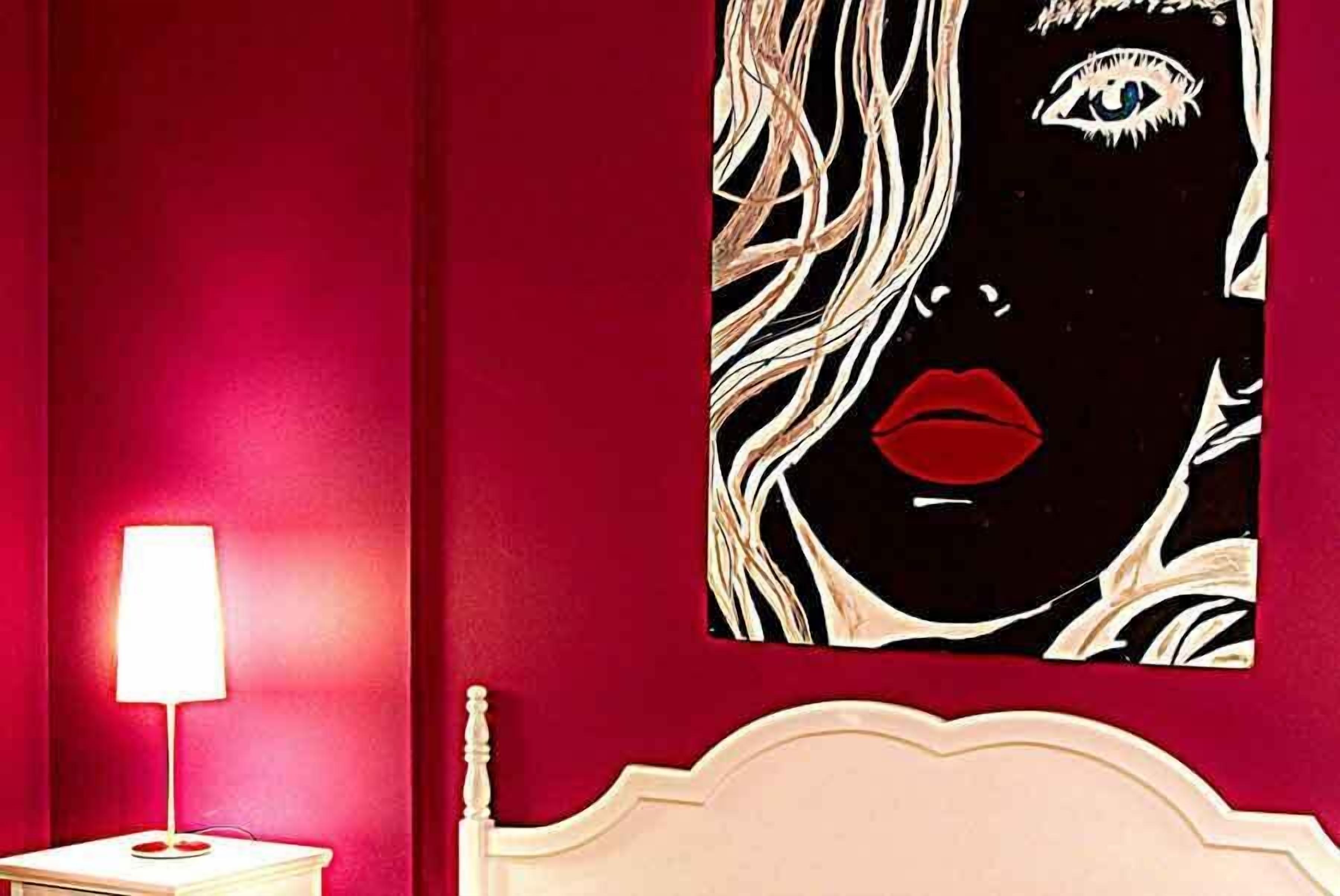 Quarto com parede vermelha, mesa de cabeceira branca com abajur e uma grande obra de arte emoldurada acima da cama.
