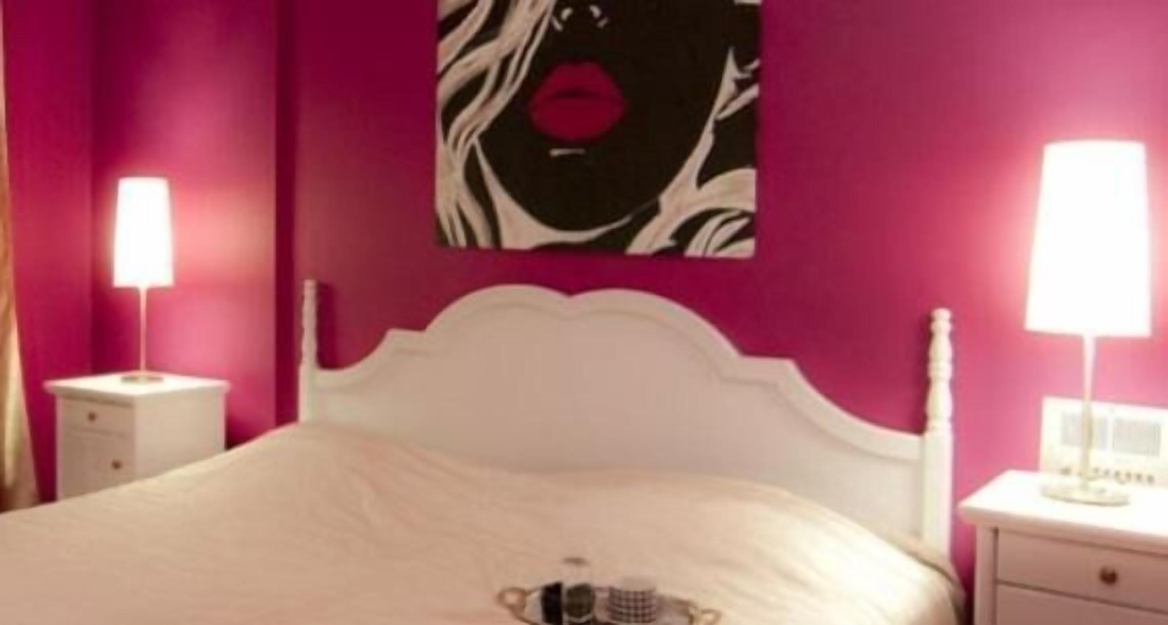 A bedroom with a white bed, pink walls, and a painting of lips. There are two bedside tables with lamps.