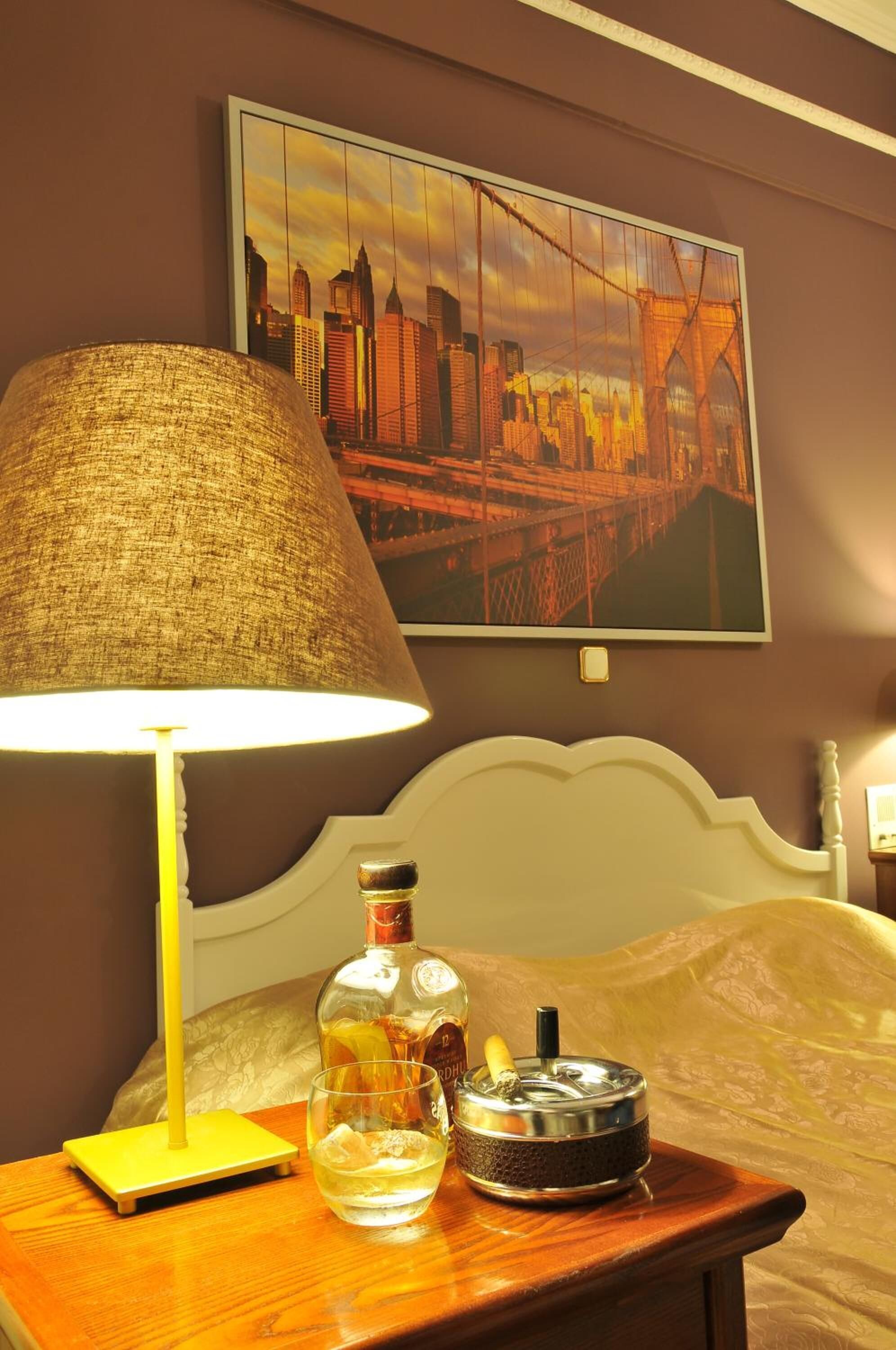 Quarto de hotel com uma cama, mesa de cabeceira, abajur, garrafa e cinzeiro.