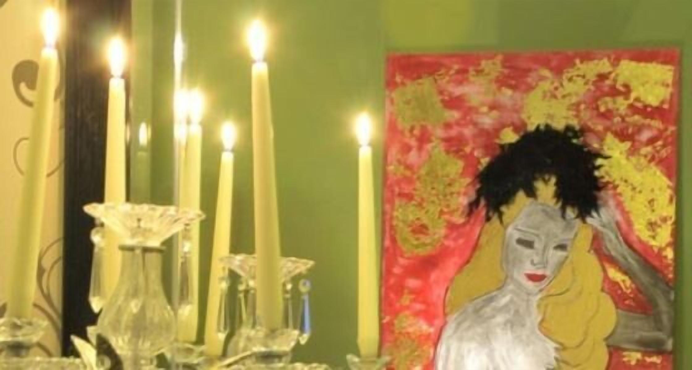 Una habitación con pared verde, un cuadro con una figura y varias velas encendidas.