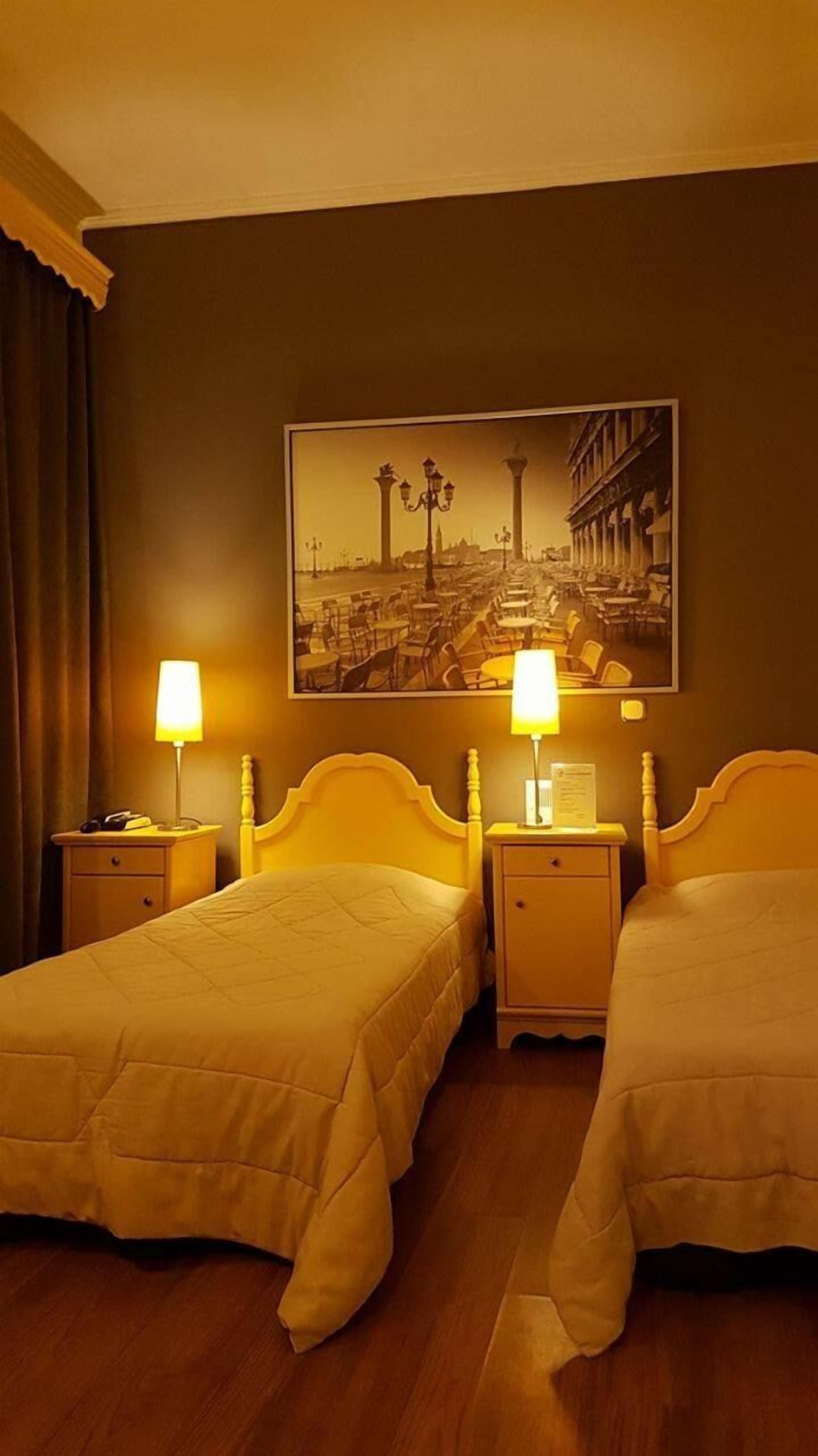 Habitación de hotel con dos camas, una mesita de noche con lámpara, un cuadro enmarcado en la pared y suelo de madera.