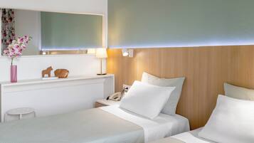 A hotel room with two beds, a bedside table with a lamp, a mirror, and a small shelf with decorative items.