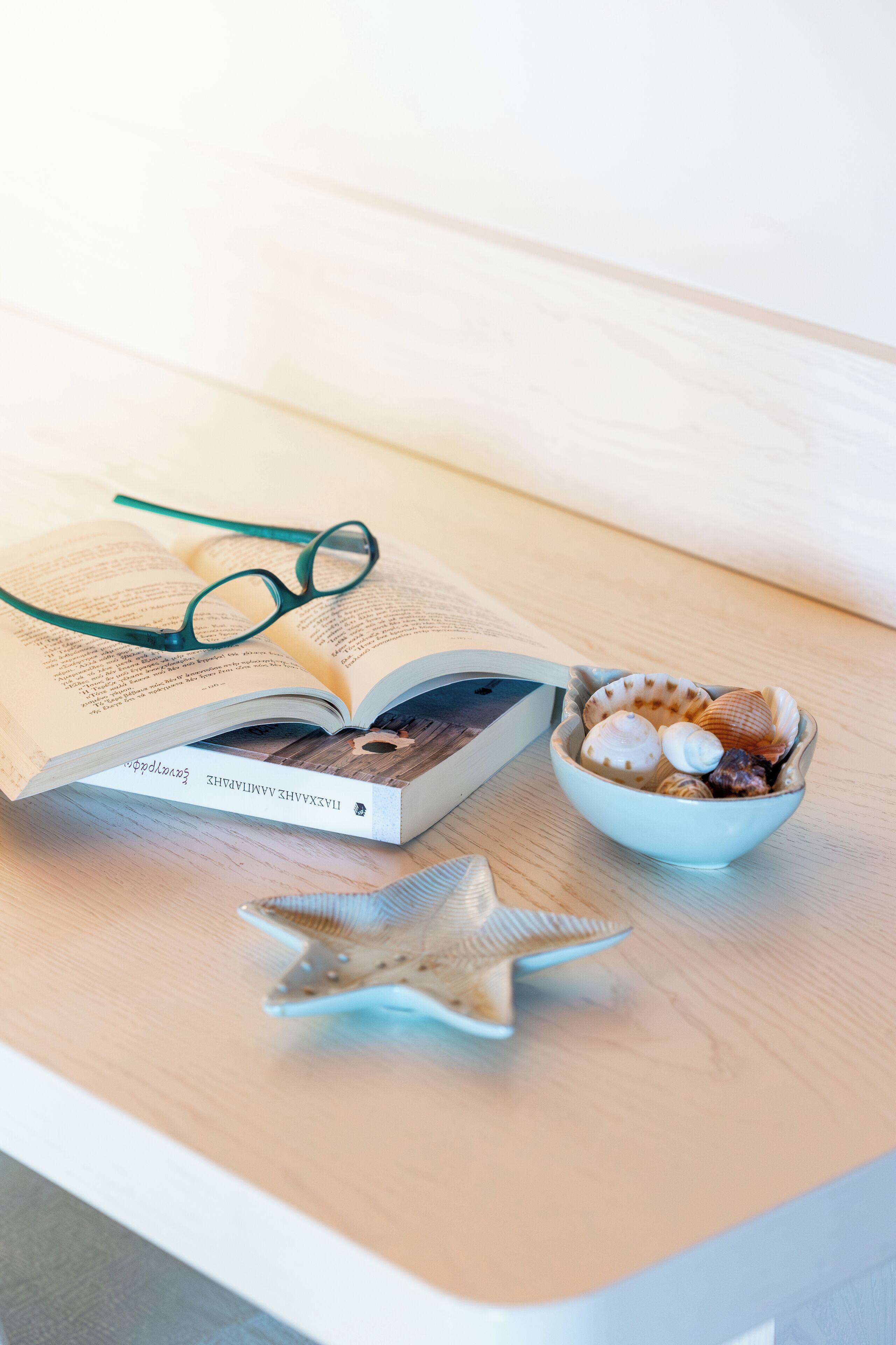 A book, a pair of glasses, a bowl of seashells, and a starfish on a wooden surface.