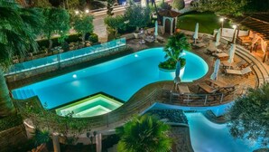A large, curved swimming pool with a glass bottom, surrounded by lounge chairs and umbrellas, and a small hot tub. - Hotel Pyrgos Boutique & Suites (Aristotelis)