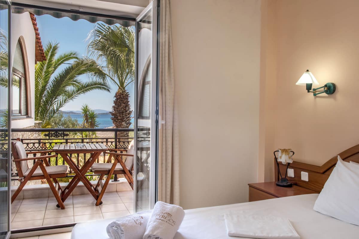 A hotel room with a bed, a nightstand, a lamp, a window with a view of palm trees and the sea, and a balcony with a table and chairs.