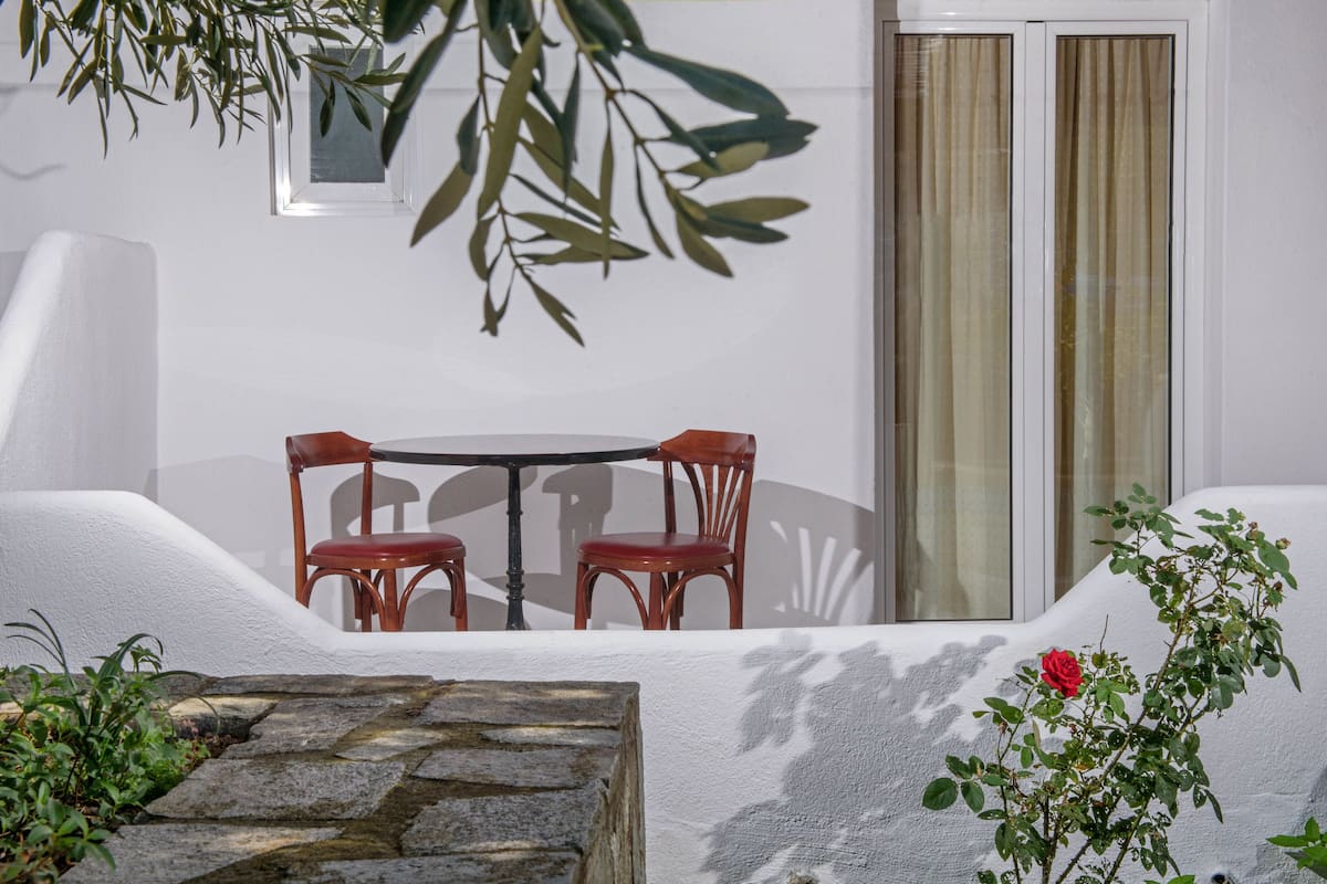 A white-walled room with a round table and two chairs, a window with curtains, and a potted plant.
