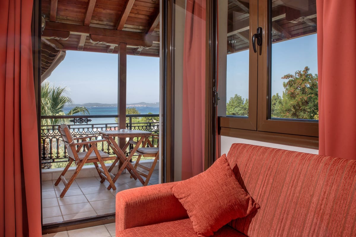 A balcony with a wooden table and chairs, a red sofa, and a view of the sea and hills.