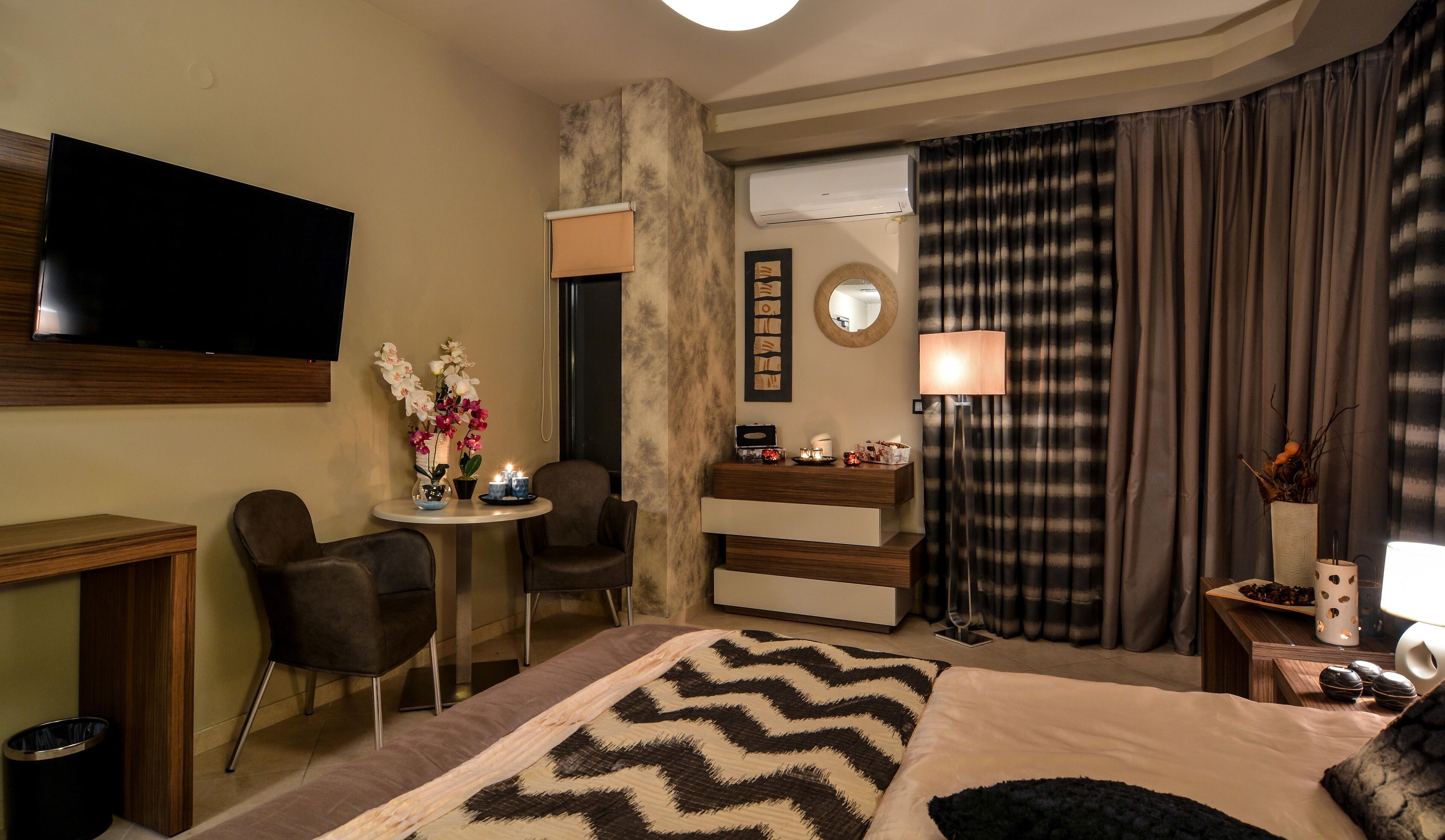 A hotel room with a bed, a TV, a desk with a chair, a small table with candles, a mirror, and a lamp.