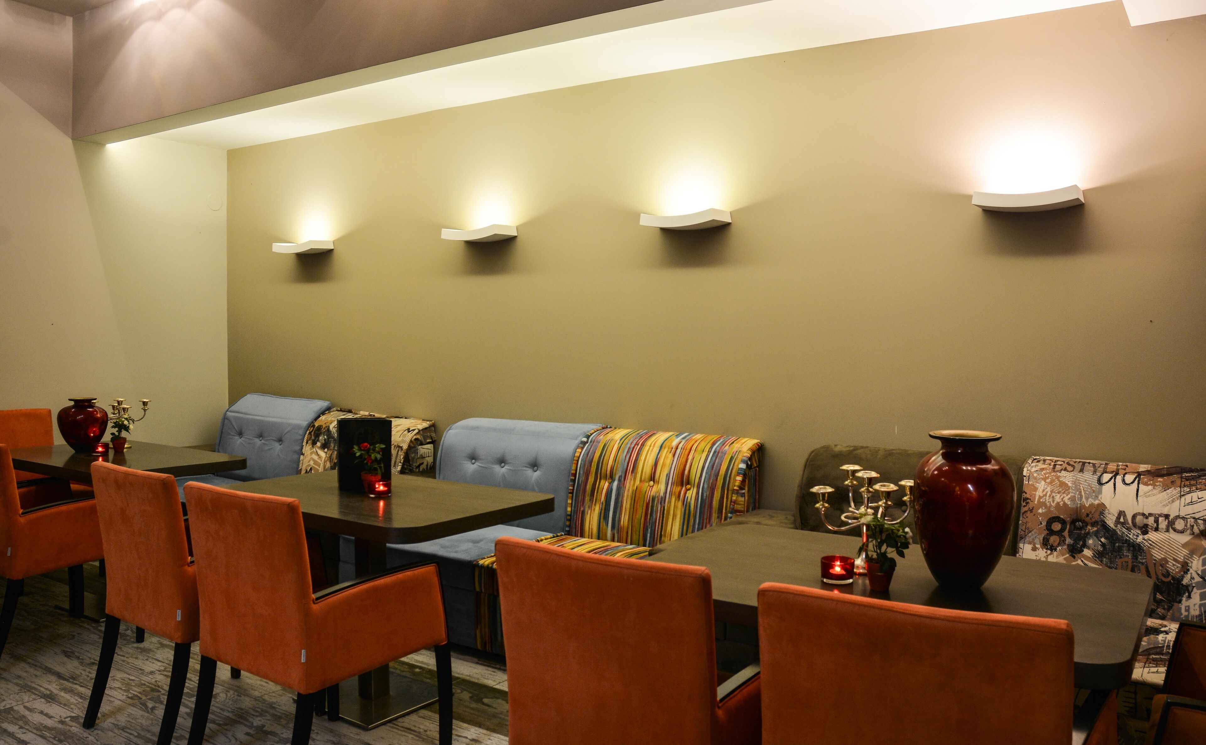 Uma sala de jantar com uma mesa comprida, cadeiras laranja, um sofá com um cobertor colorido e luminárias fixadas na parede.