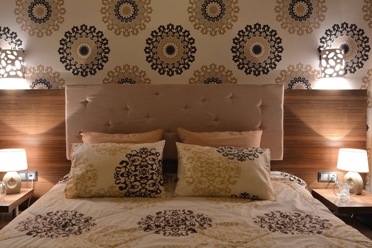 Uma cama bem arrumada com colcha estampada, duas almofadas iguais e cabeceira acolchoada. Há duas mesas de cabeceira com abajures e um relógio de parede.