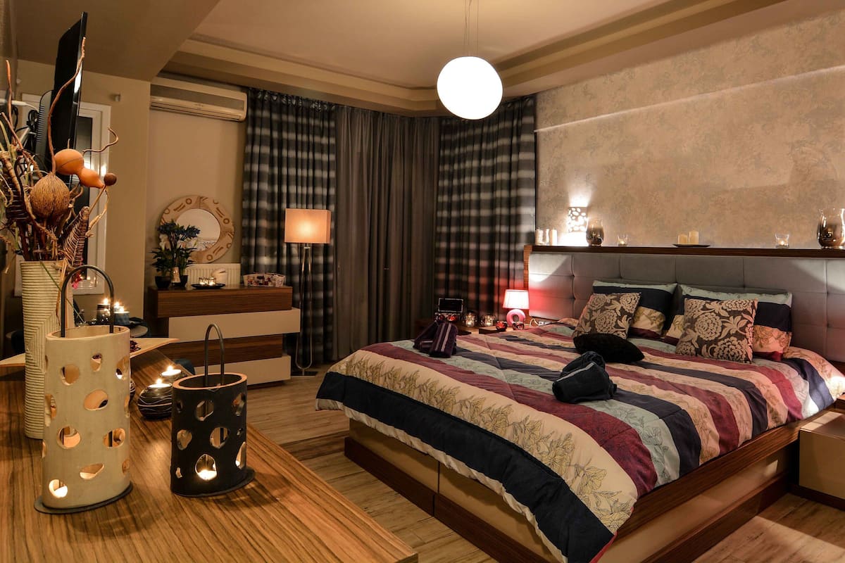 Um quarto com cama, mesas de cabeceira, luminária, espelho, cômoda e um vaso com flores.
