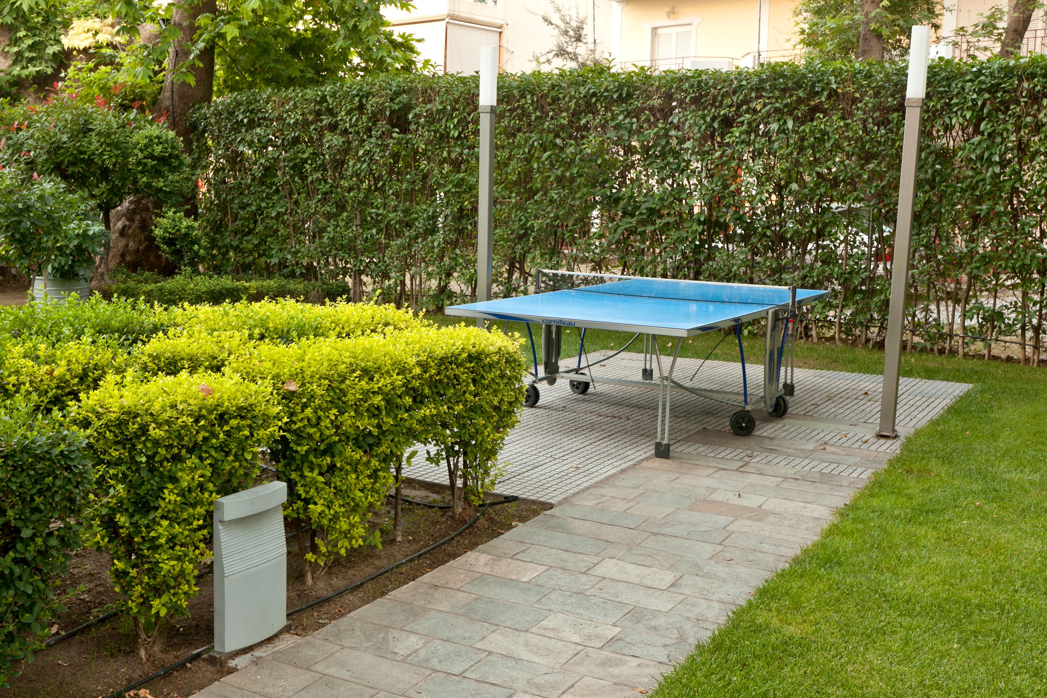 A ping pong table in a garden area with a paved walkway, surrounded by well-maintained hedges and a building in the background.