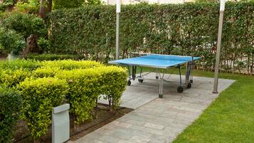 A ping pong table in a garden area with a paved walkway, surrounded by well-maintained hedges and a building in the background.