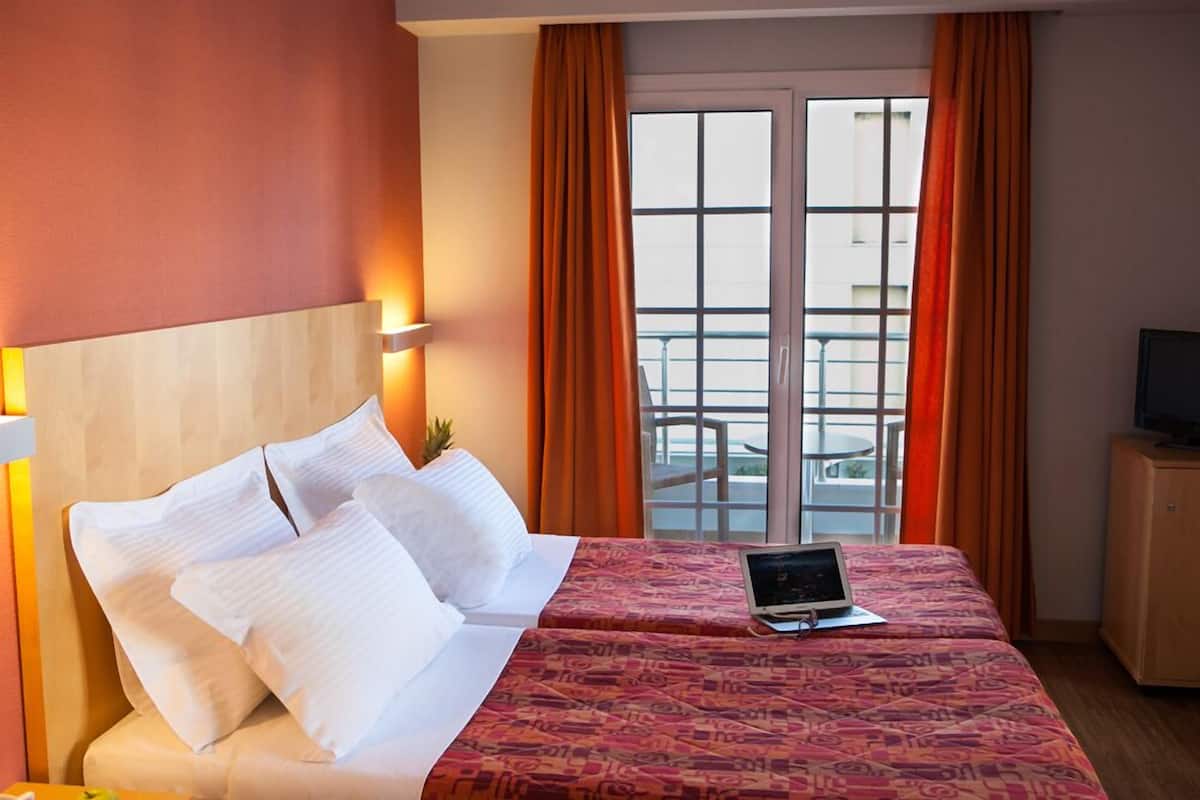 A hotel room with a bed, a laptop on the bed, a TV on a cabinet, and a balcony with orange curtains.