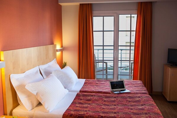Habitación de hotel con cama, una laptop sobre la cama, un televisor en un mueble y un balcón con cortinas naranjas.