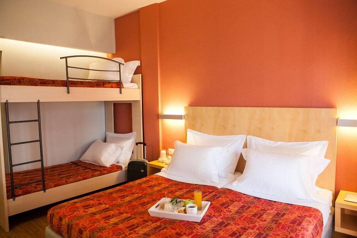 A hotel room with two bunk beds, a wooden headboard, a bedside table with a lamp, and a tray with breakfast items.