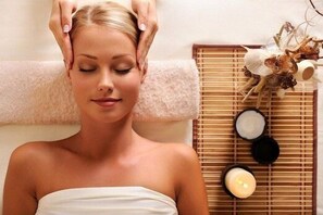 Une personne reçoit un massage de la tête, enveloppée dans une serviette, avec une bougie et un bol de sel placés sur un tapis en bambou.