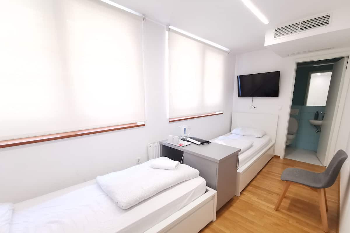 Une chambre d’hôtel avec deux lits simples, un bureau avec une chaise, un téléviseur fixé au mur et une salle de bain visible à travers une porte ouverte.