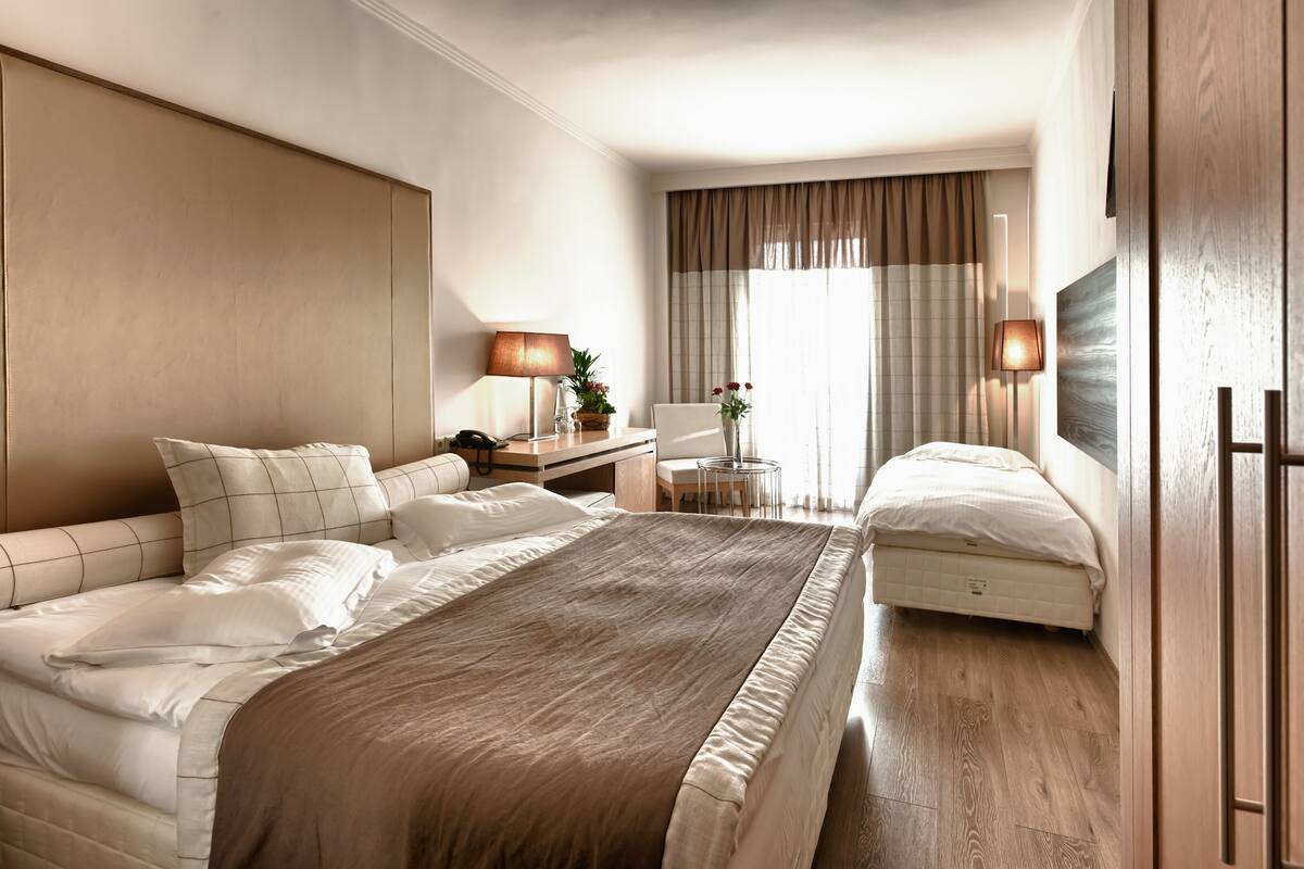 Habitación de hotel con una cama grande, mesitas de noche, un escritorio, una silla, una pequeña mesa con lámpara y un ventanal con cortinas.