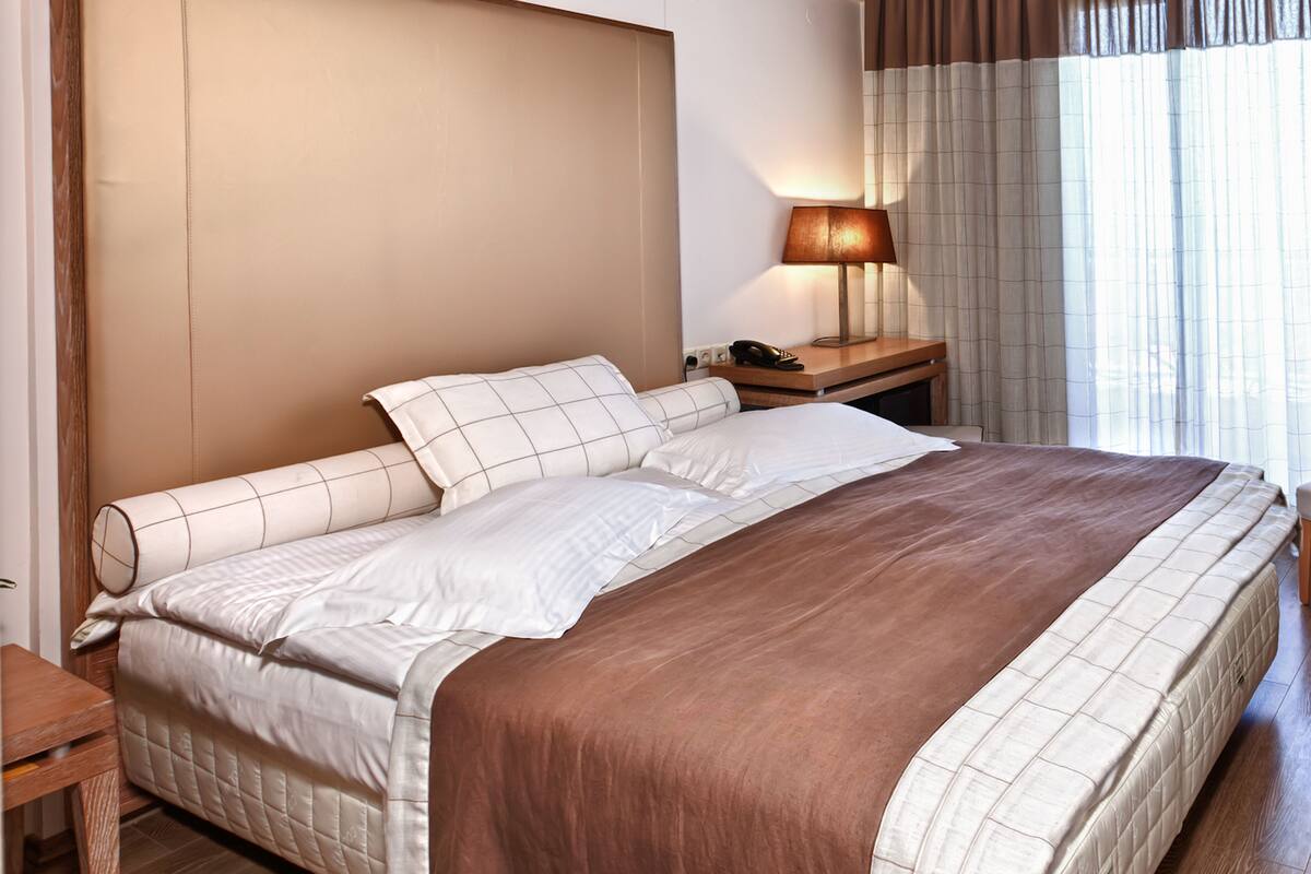 Una habitación de hotel con una cama grande, una mesita de noche con lámpara y un ventanal con cortinas.