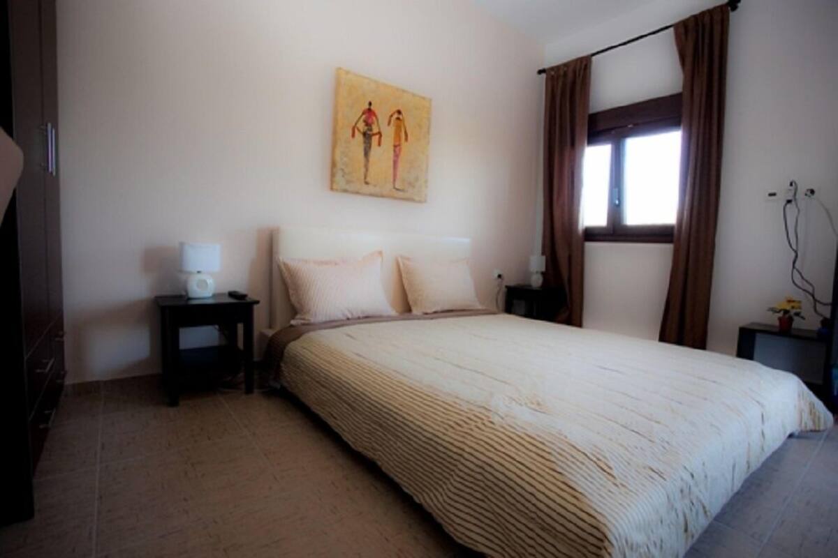 Ein Schlafzimmer mit Bett, Nachttisch, Wandbild und Fenster mit Vorhängen sowie einer Tür.