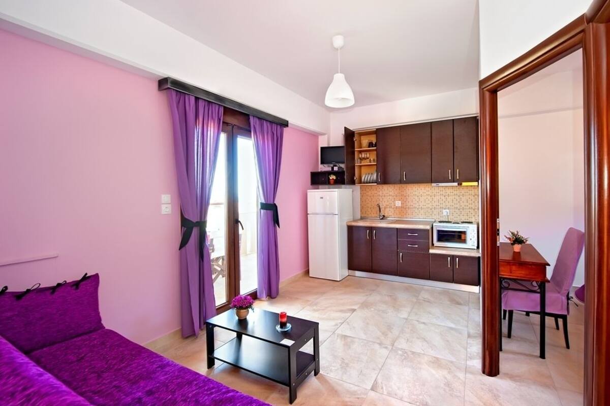 Ein kompakter Wohnbereich mit einem violetten Sofa, einem kleinen Esstisch und einer Küchenzeile im Hintergrund.