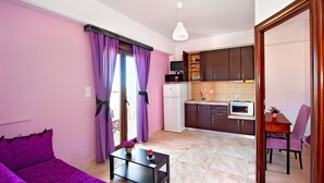 Un espacio habitable compacto con un sofá morado, una pequeña mesa de comedor y una kitchenette al fondo.