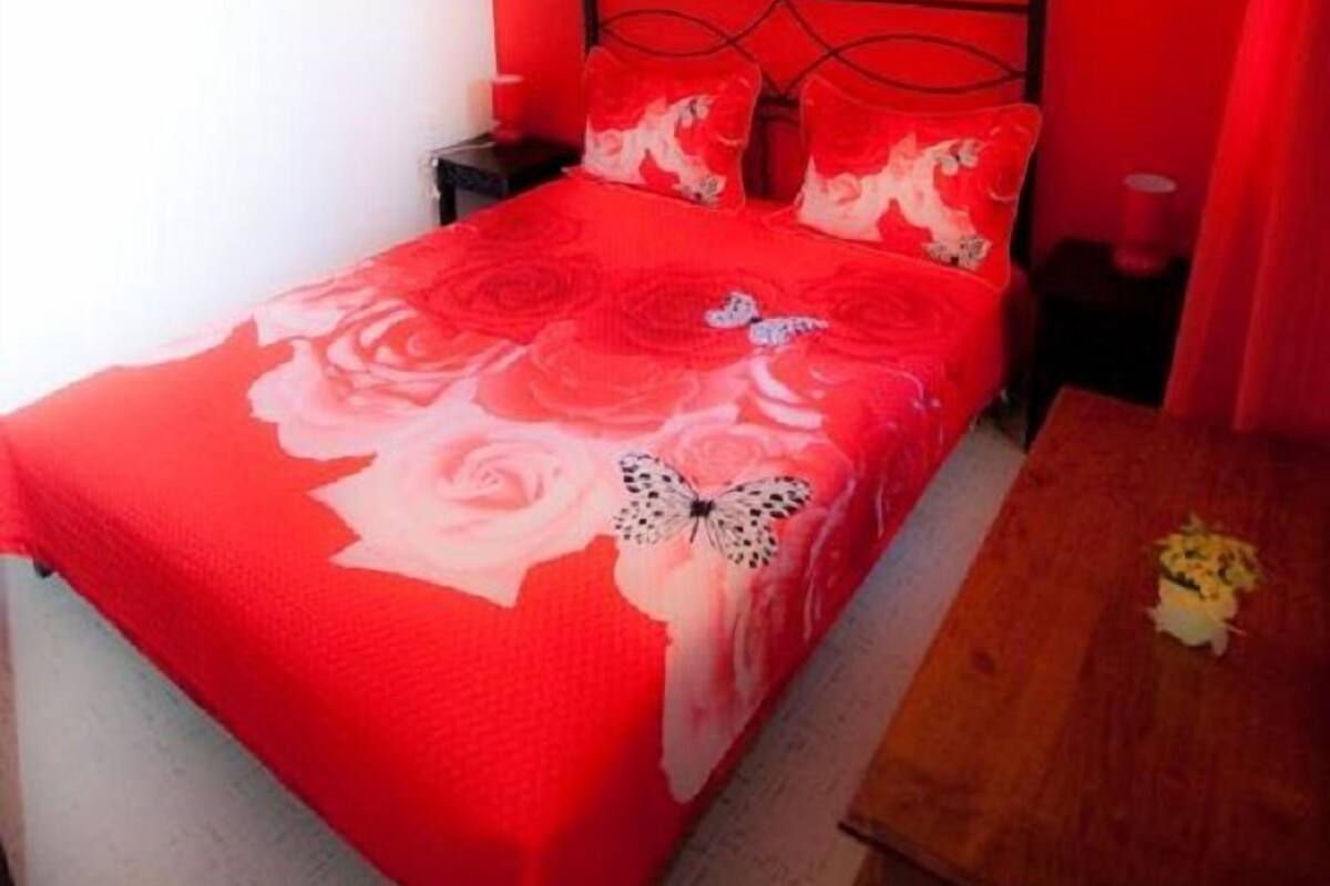 Ein Schlafzimmer mit roter Wand, einem Bett mit blumigem Überwurf, einem Nachttisch, einem Holztisch und einem Gemälde über dem Bett.