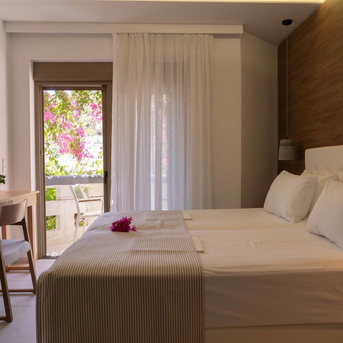 Habitación de hotel con cama, escritorio y silla. Se ve una ventana con vistas a flores rosadas y un balcón.