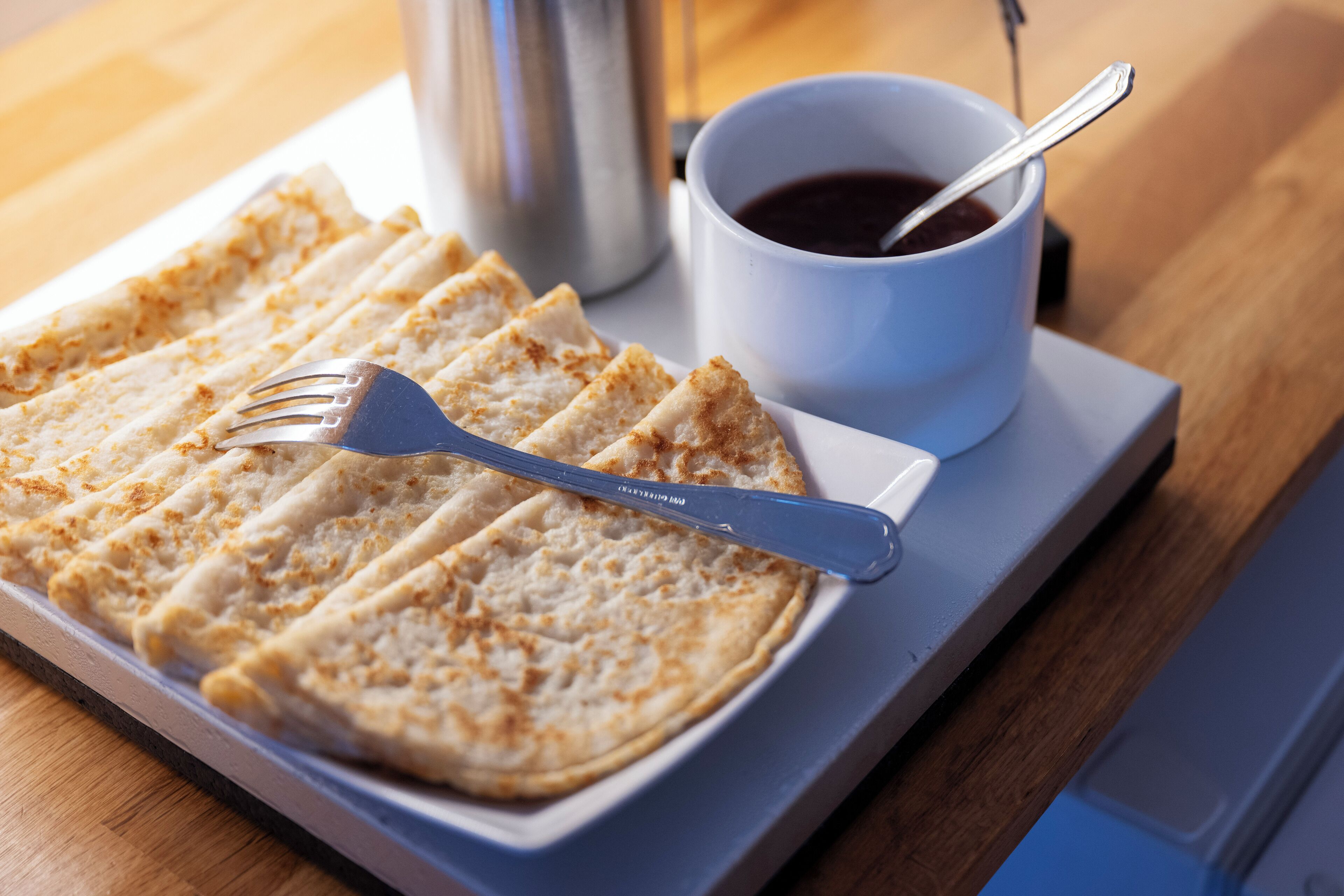 Un plato de crepas, un tenedor, una taza de café y una termo sobre una bandeja.