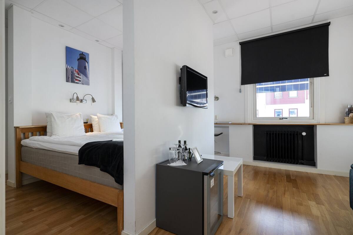 A bedroom with a bed, a desk, a TV, and a window with a view of buildings.
