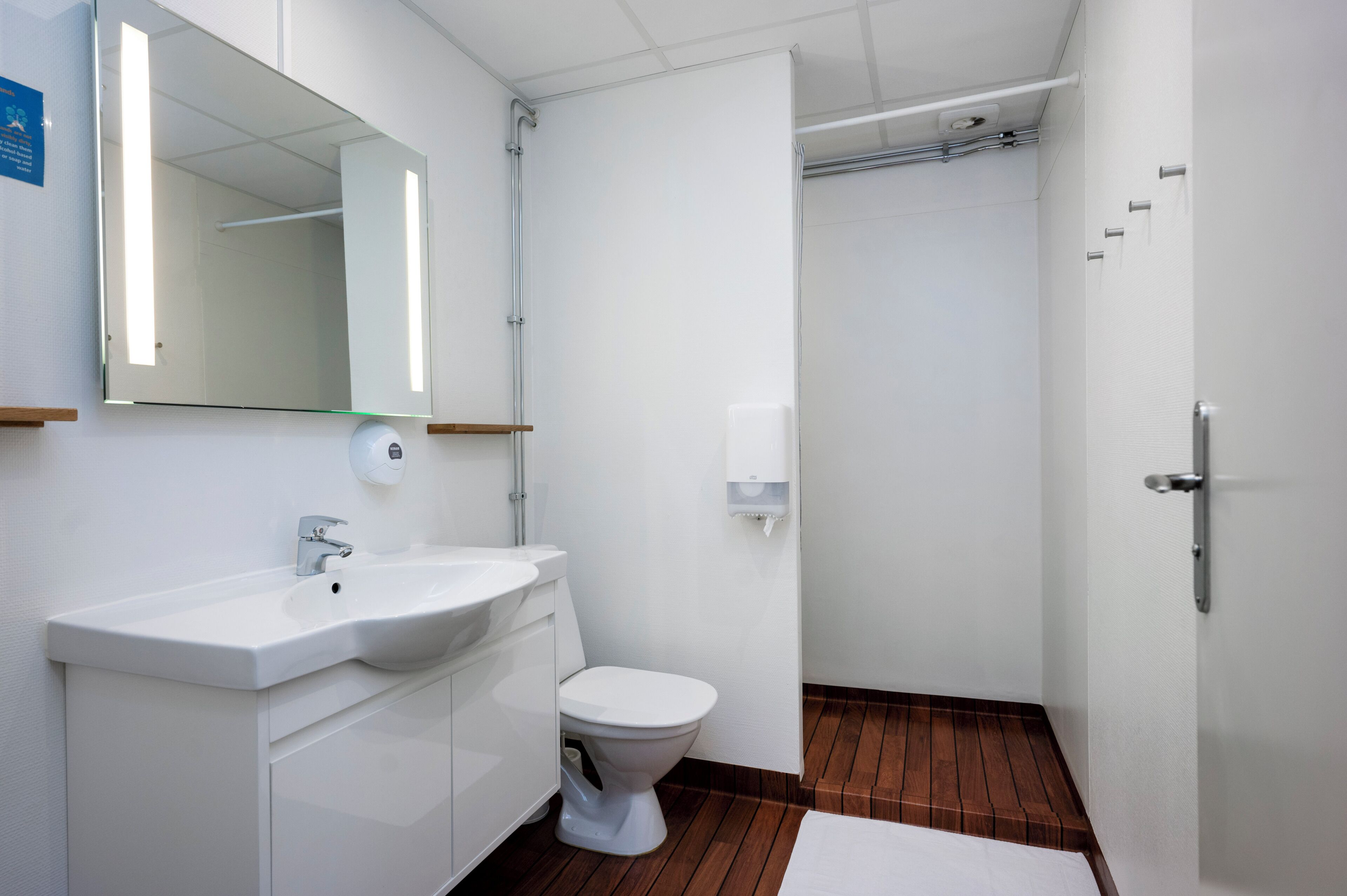 A modern bathroom with a white sink, mirror, and toilet, wooden flooring, and a white towel on the floor.