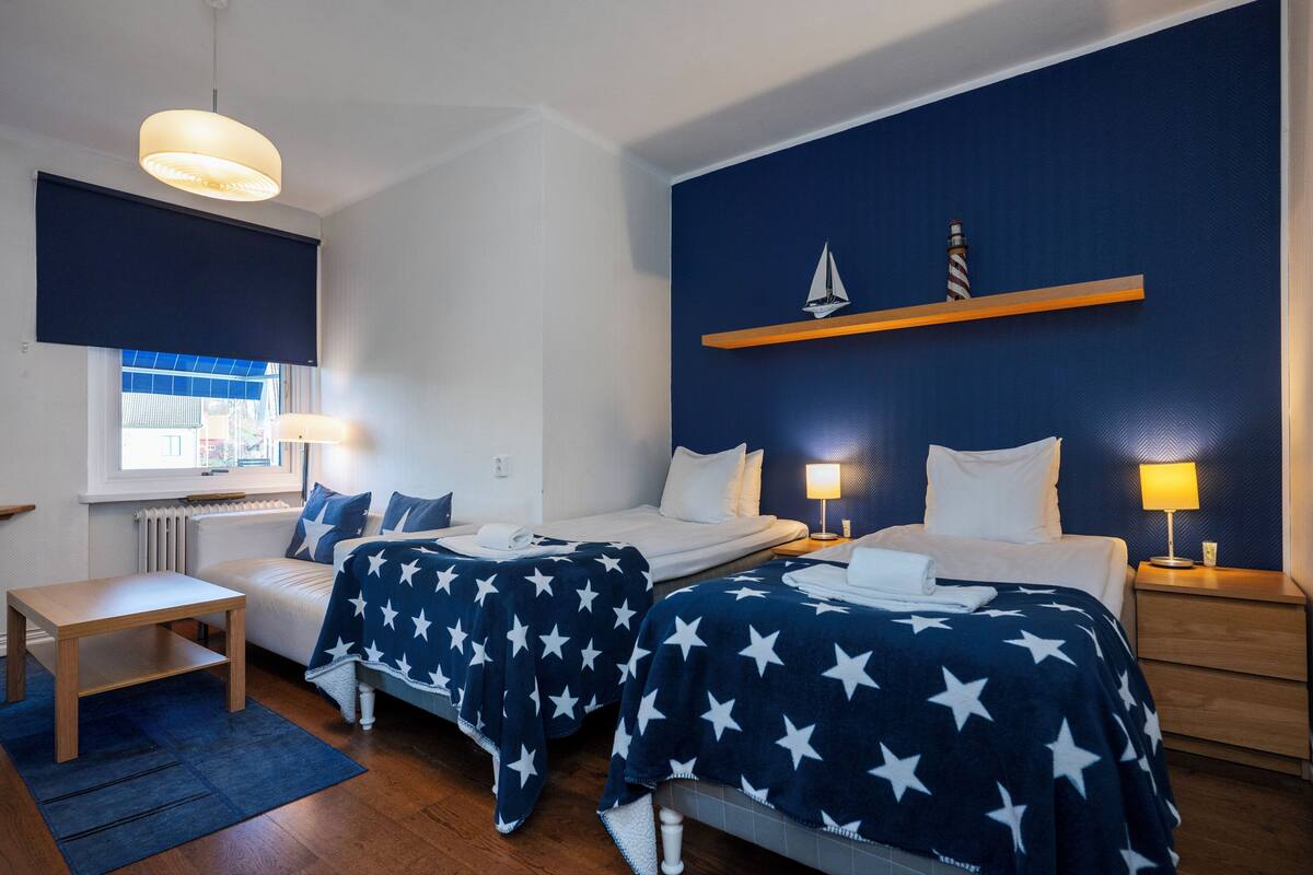 A hotel room with two beds, a small table, a chair, a window with blinds, a radiator, and a shelf with a model ship.