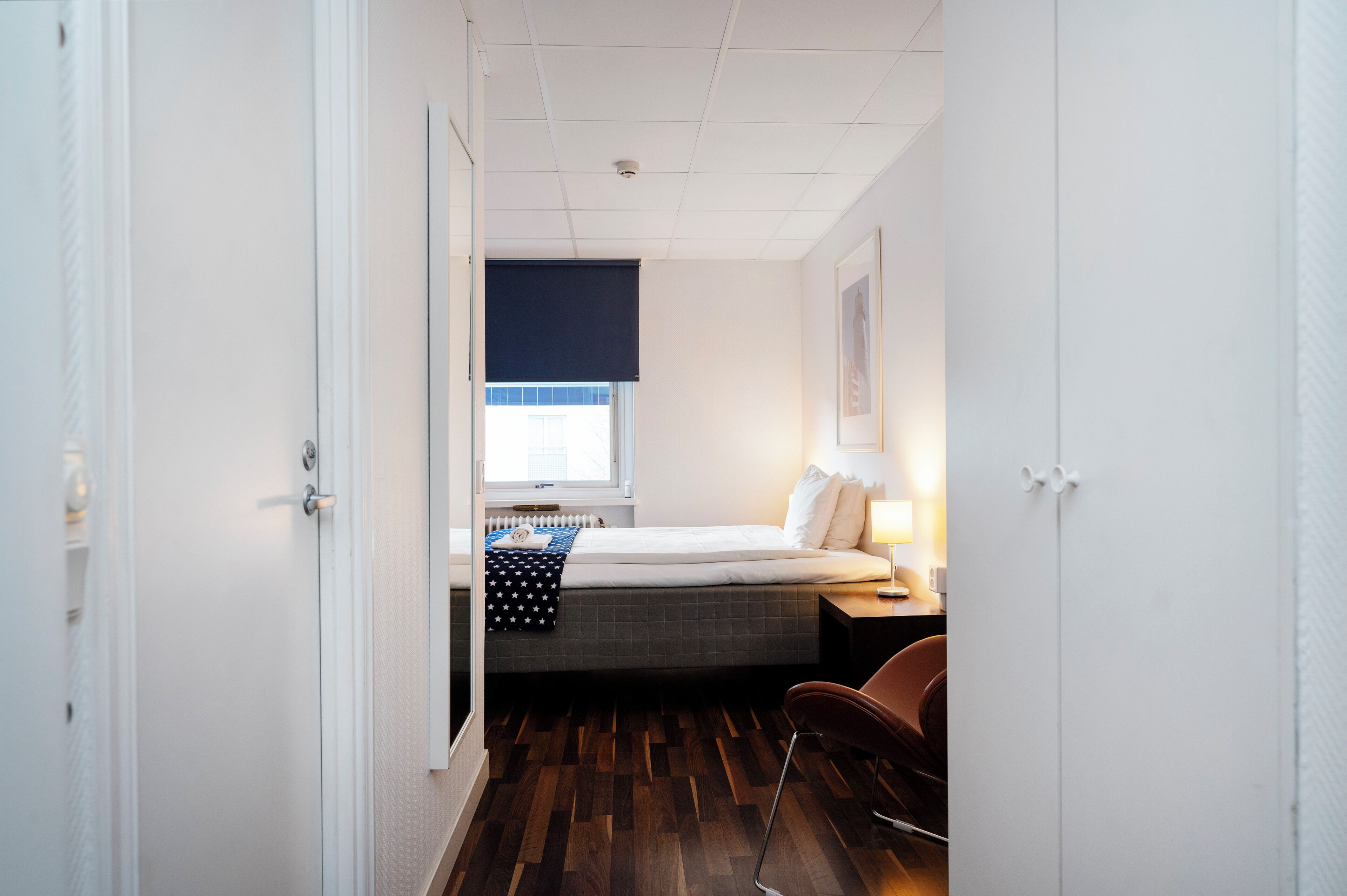 A small, neatly arranged bedroom with a bed, bedside table, lamp, and a window with a view.