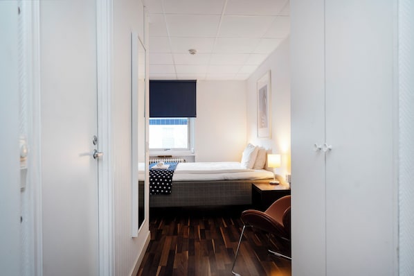A small, neatly arranged bedroom with a bed, bedside table, lamp, and a window with a view. - Port Hotel (Karlshamn)