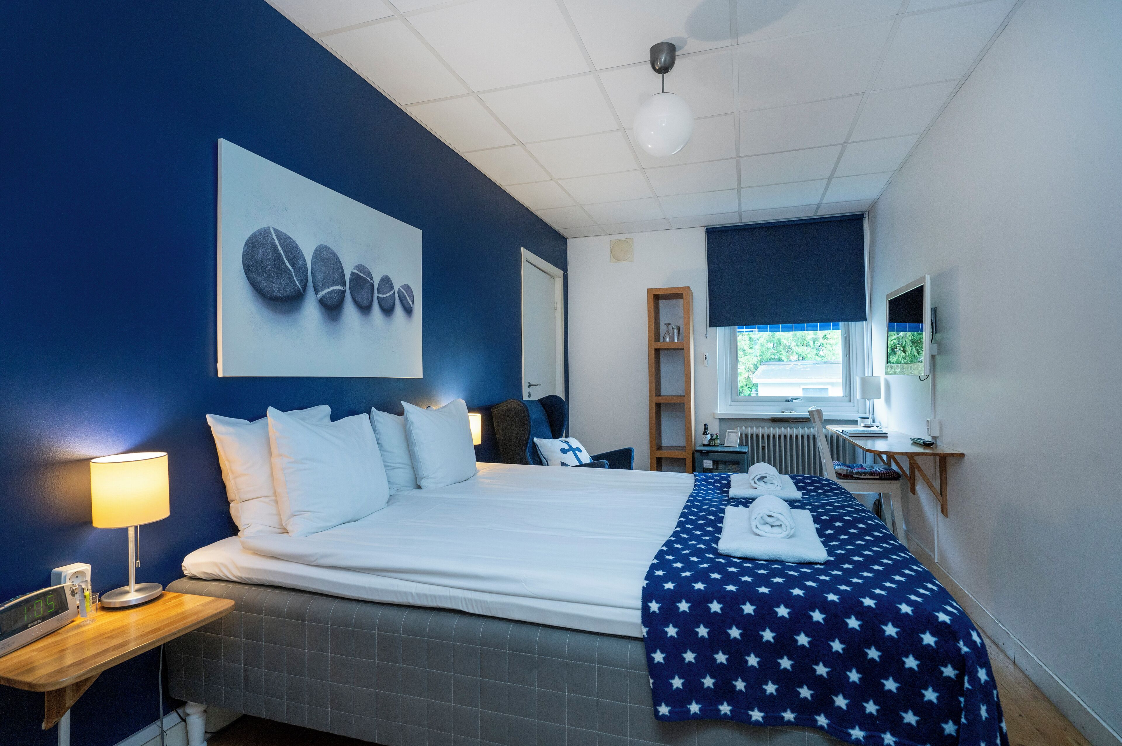 A hotel room with a bed, a desk, a lamp, a clock, and a window.