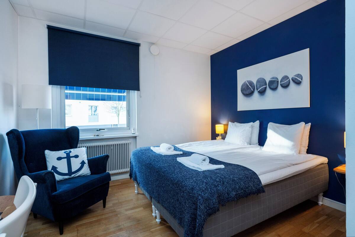 A hotel room with a bed, a chair, a window, and a wall with a painting.