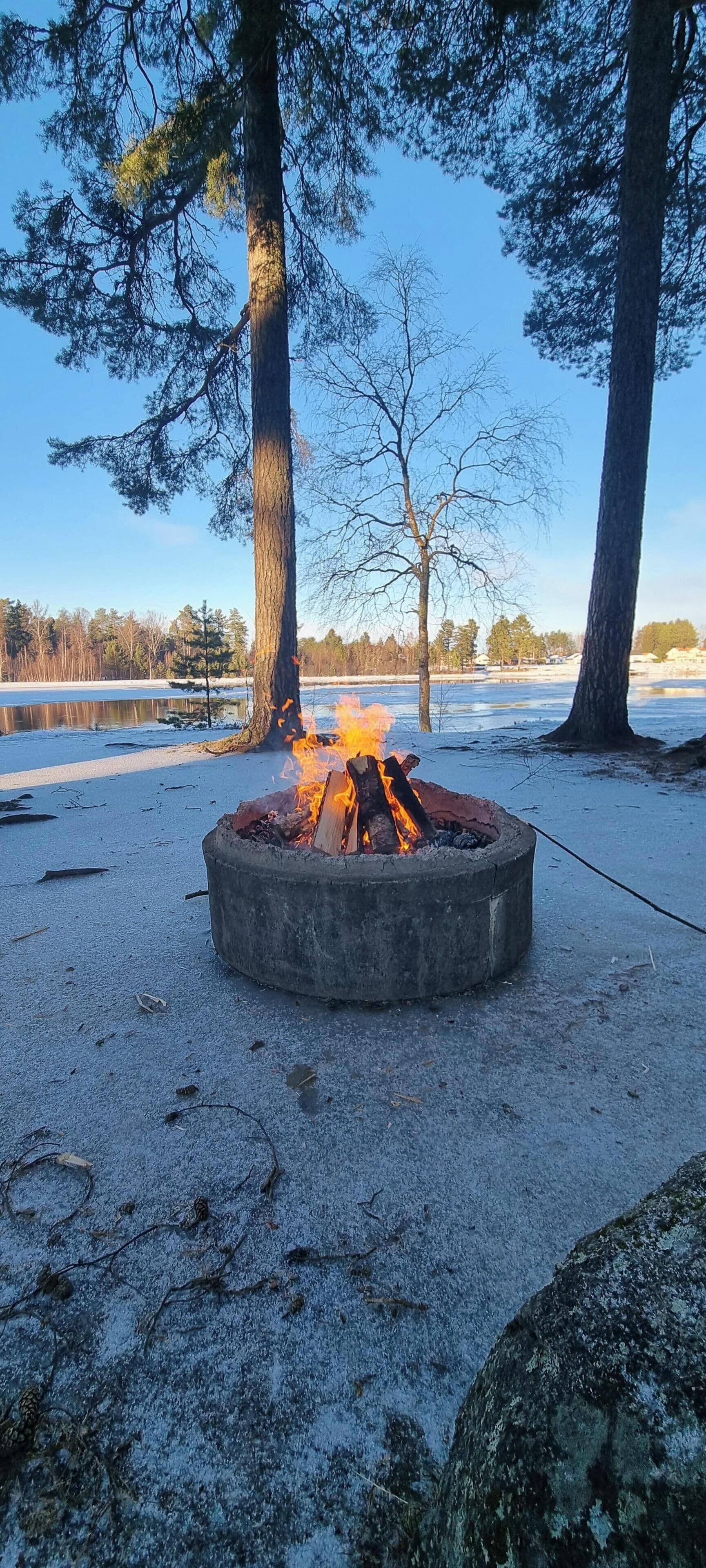 A concrete fire pit with flames, surrounded by snow and trees, with a clear sky in the background.
