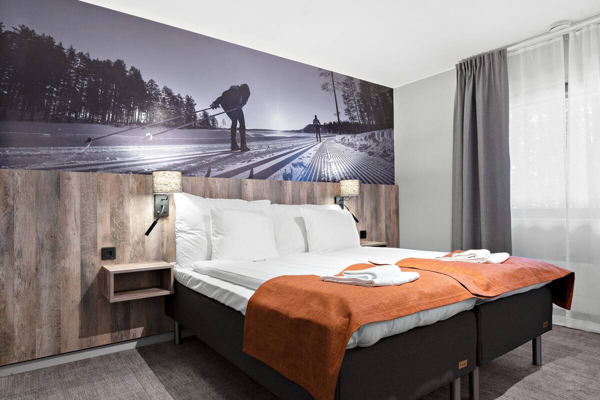 A hotel room with a large bed, a wooden headboard, a nightstand, a wall-mounted lamp, and a large window with curtains.