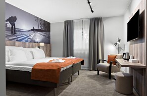 A modern hotel room with a large bed, a desk with a chair, a flat-screen TV, and a large window with curtains. - First Camp Moraparken (Mora)