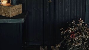 Interior escuro de madeira com castiçal de chifres e velas, uma planta em vaso e um cesto de flores secas.