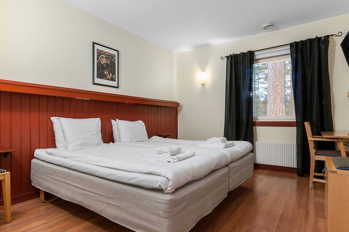 A neatly made bed with white linens, a wooden headboard, a small table with a chair, and a window with curtains.