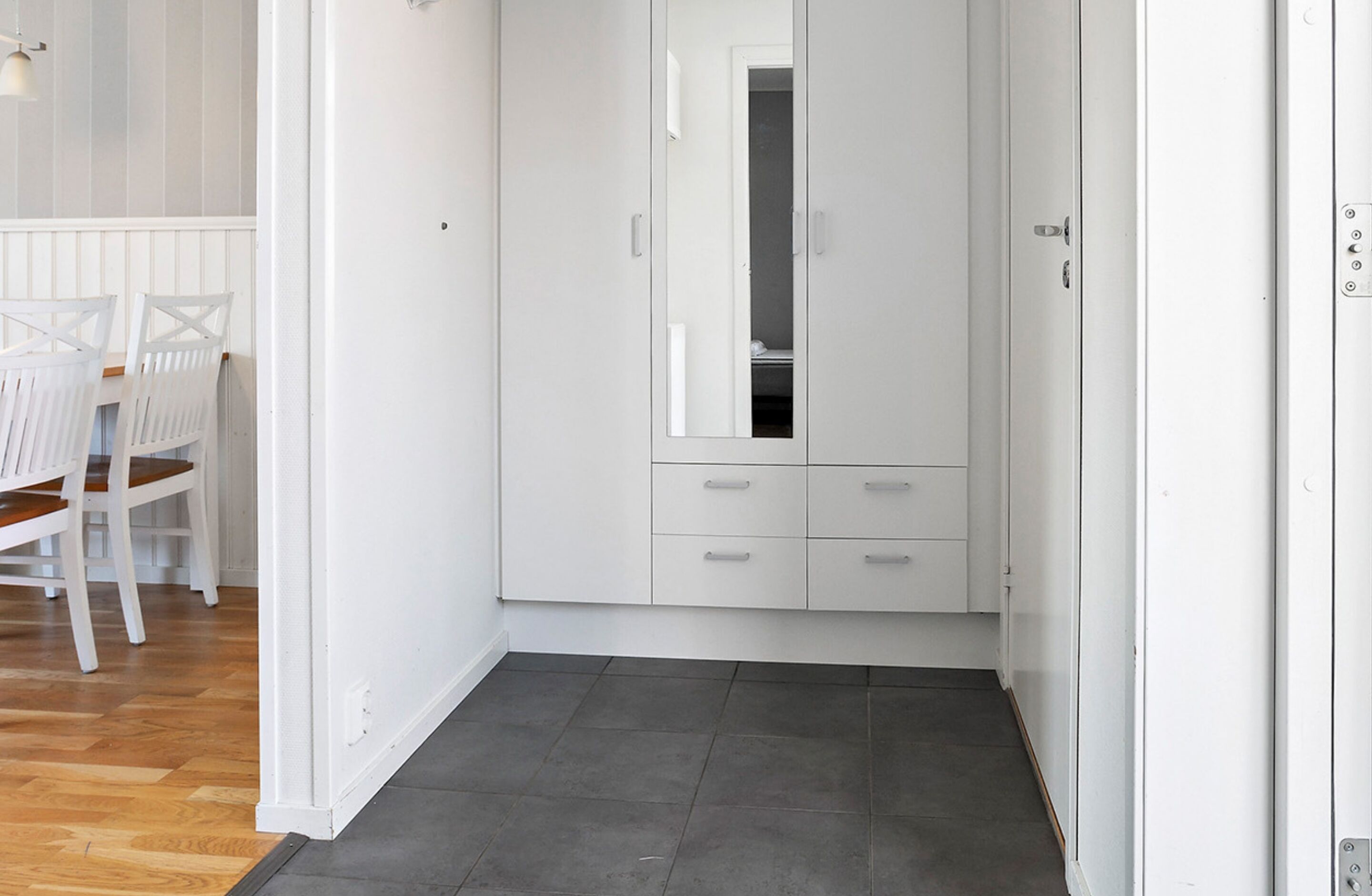 A white room with a wooden floor, a white wardrobe with drawers, and a mirror.