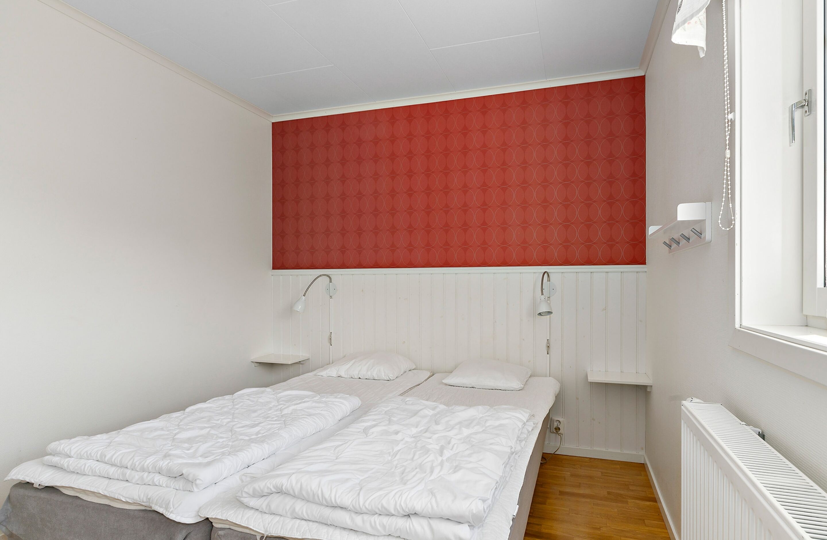 Une chambre à coucher avec un lit, deux tables de chevet, un mur d’accent rouge et une fenêtre avec un rideau blanc.