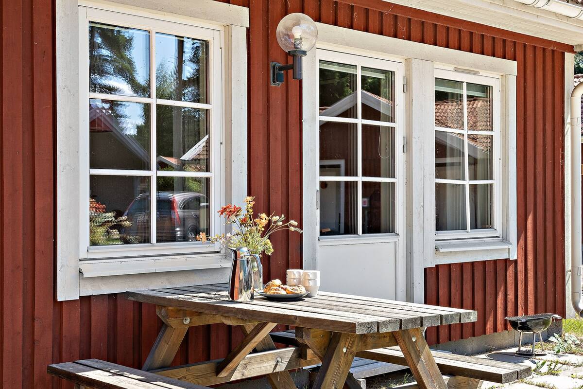 A wooden picnic table with a vase of flowers and a pitcher, set against a red wooden wall with two white-framed windows.