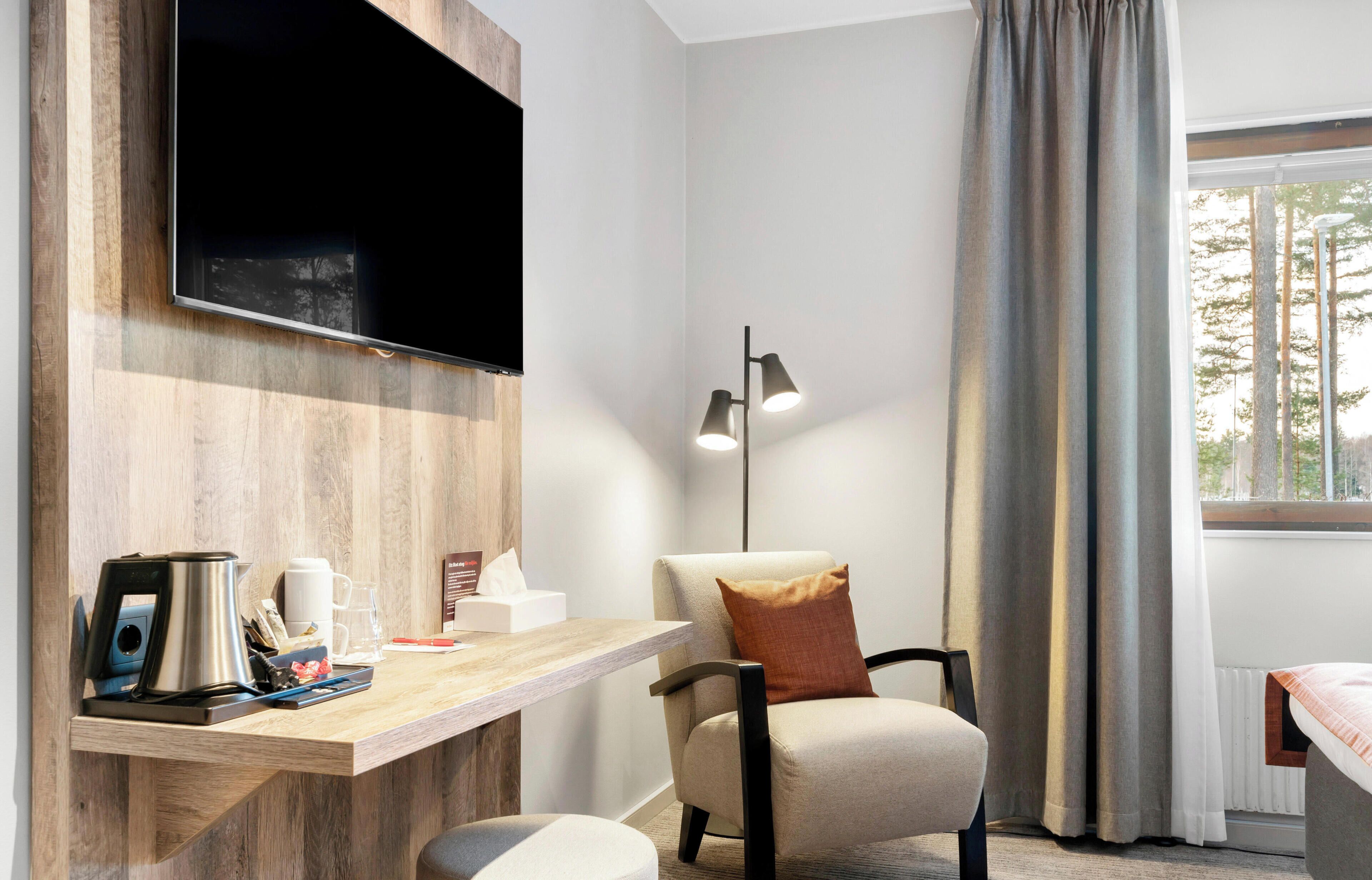 Una habitación moderna con una televisión de pantalla plana, un estante de madera con tetera y cafetera, una silla con cojín, una lámpara de pie y una ventana con cortinas.