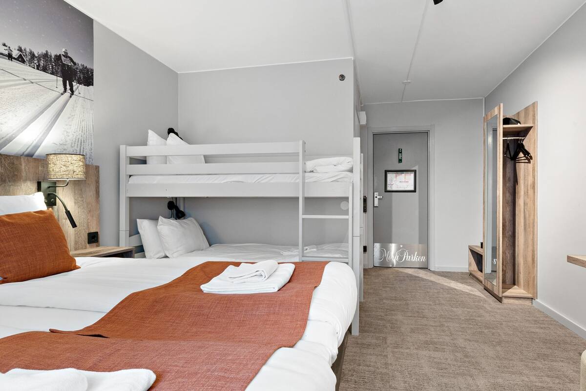 A hotel room with a bunk bed, a single bed, a wooden nightstand with a lamp, a mirror, and a door leading to another room.