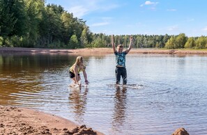 Two people standing in shallow water near a sandy beach, with trees and a clear sky in the background. - First Camp Moraparken (Mora)