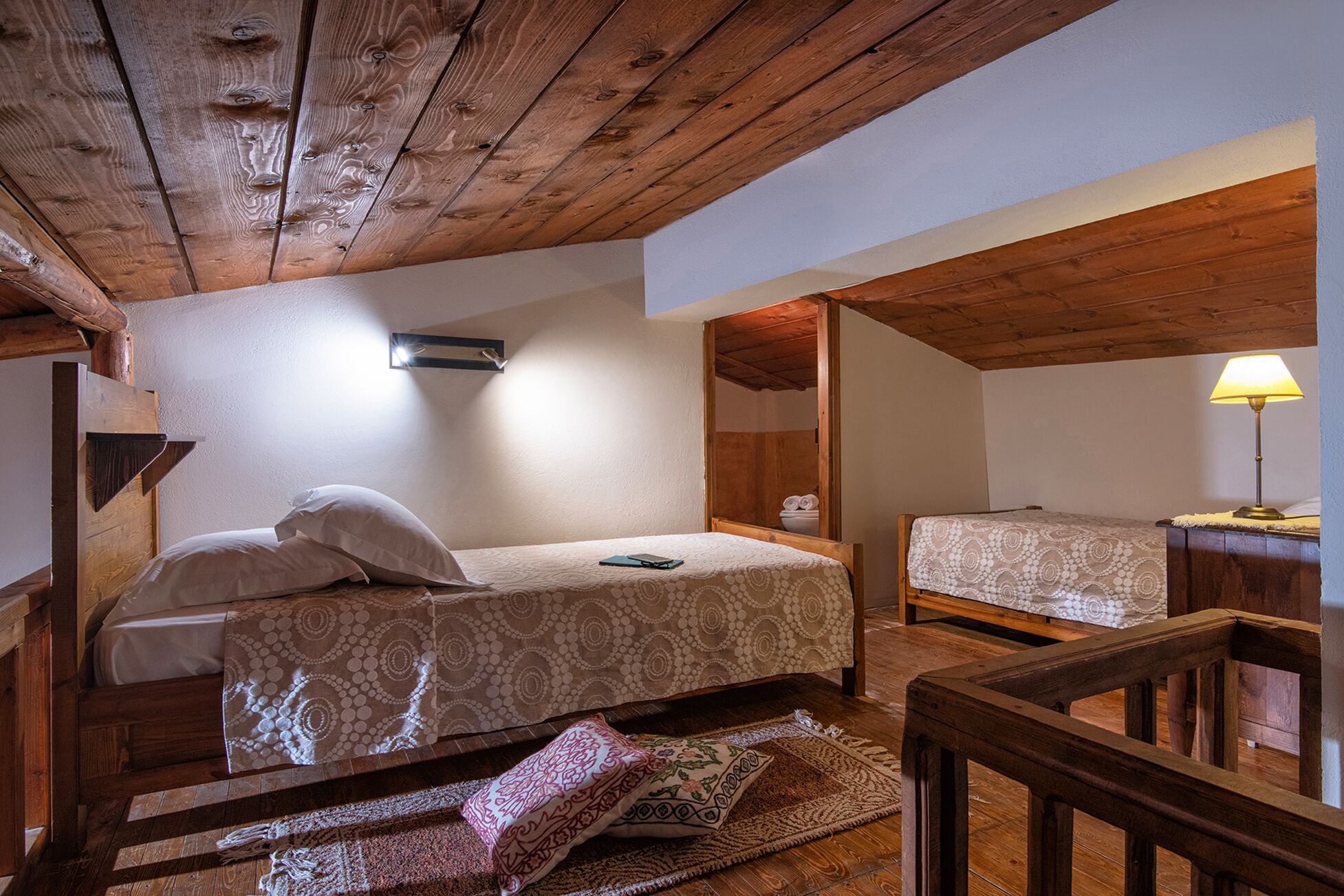 A cozy attic bedroom with a wooden ceiling, a single bed with a patterned bedspread, a bedside table with a lamp, and a small bathroom visible through an open door.